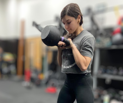 A woman is holding a heavy kettlebell in a gym, demonstrating strength and proper form, which are key components for individuals pursuing or benefiting from personal trainer certification.