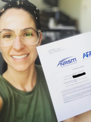 A smiling woman with clear framed glasses and a bun in her hair holds up a personal trainer certification document from NASM and AFAA. The document shows 