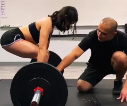 A male personal trainer is guiding a female client through a barbell deadlift, adjusting her form for safety and effectiveness, a crucial aspect of personal trainer certification.